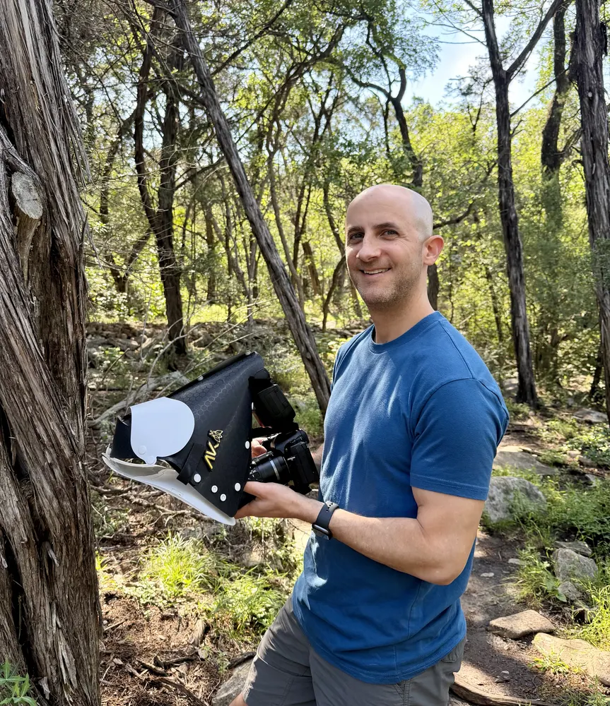Ryan smiling on a wooded trail, holding a macro camera setup with a circular diffuser.